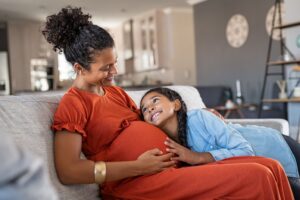 Pregnant woman in orange dress hugging her daughter on the couch